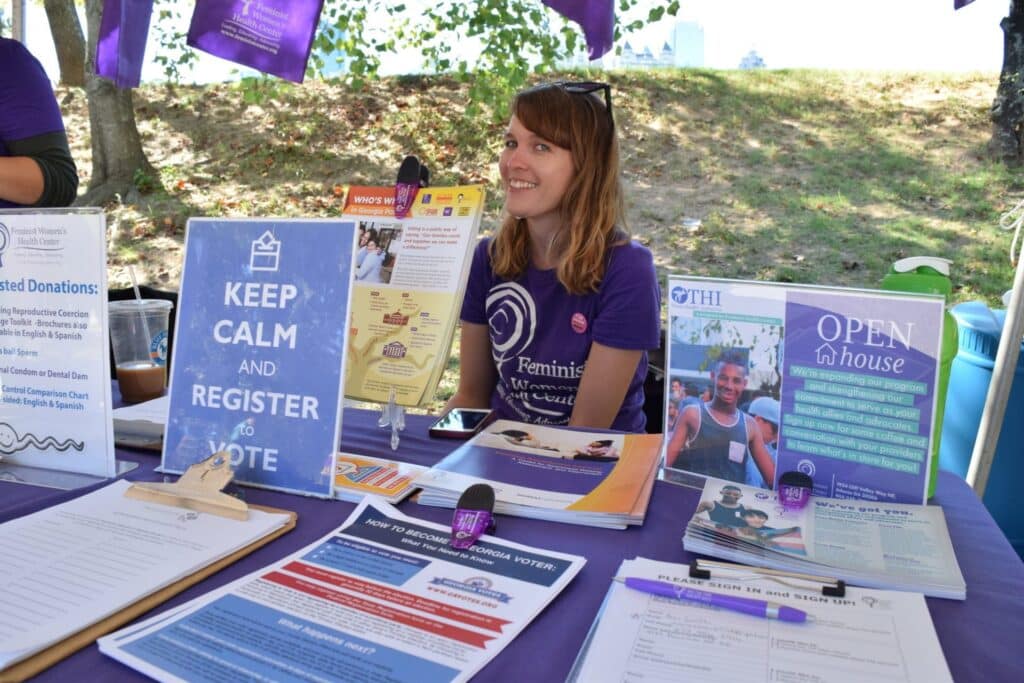 A clinic staff member smiles for a photo at an outdoor Feminist Women’s Health Center event table.