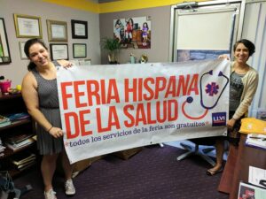 Two volunteers standing in an office holding up a banner that says "Feria Hispana de la Salud"