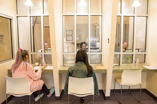 Two models pose as patients at the clinic’s front desk with three clinic staffers sitting on the other side of the glass.