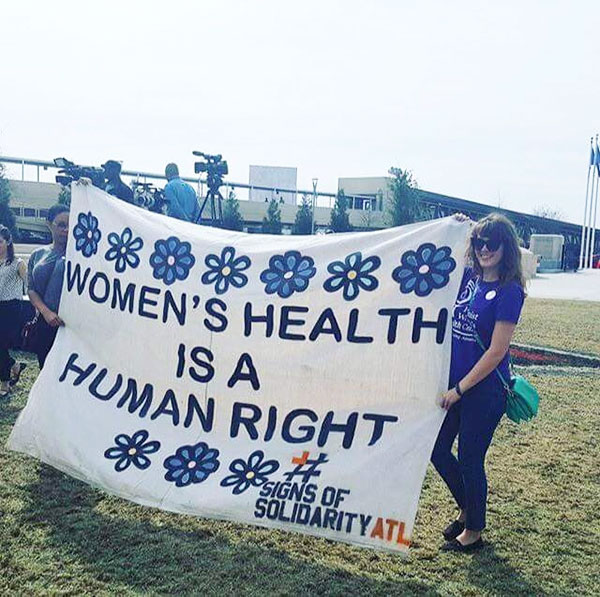 Two clinic staff members stand outside, holding a quilt that reads “Women’s Health is a Human Right.”