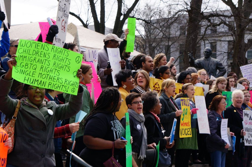 Demonstrators standing together holding signs and talking at an outdoor rally.