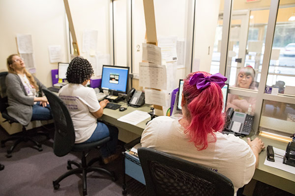 Three clinic staff members sit at the Feminist Women’s Health Center front desk, one of whom sits across from a model posing as a patient.