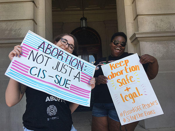 Two demonstrators pose with posters reading “Abortion is not a CIS-SUE” and “Keep Abortion Safe and Legal #PissedOffPeaches #StopTheBans” at a rally.