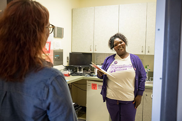 A clinic staffer smiles at a model posing at a patient with their back to the camera.