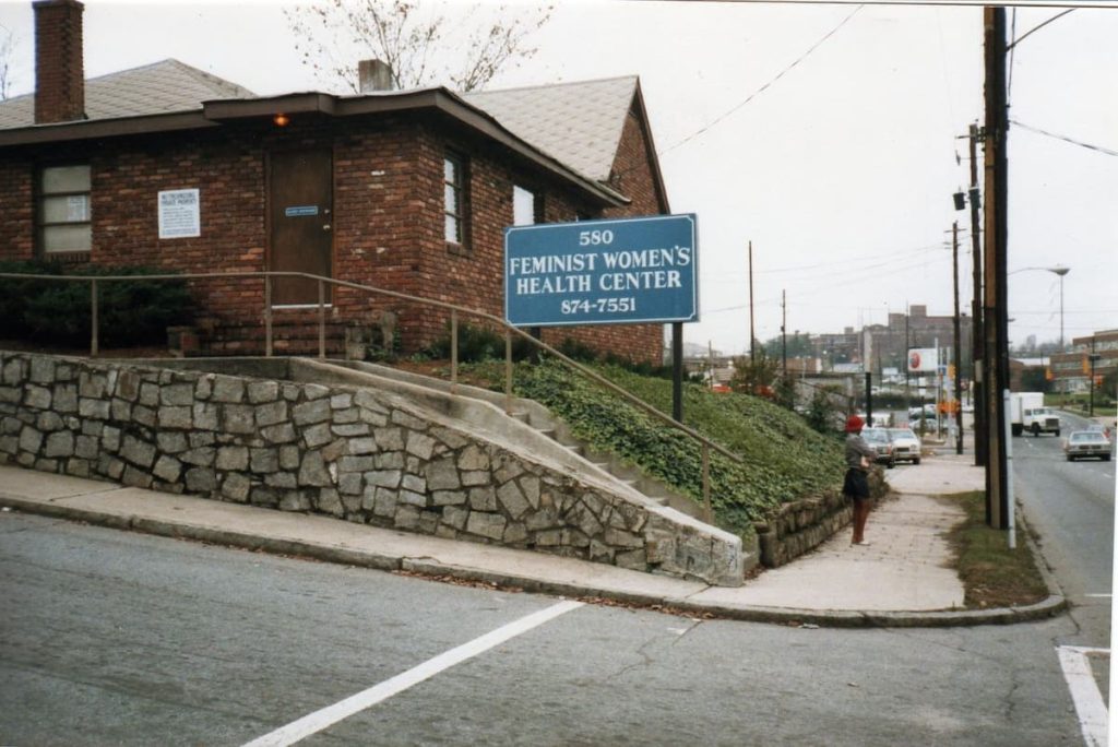 Brick clinic building on a sloped lot with a blue sign reading '580 Feminist Women's Health Center' 874-7551 beside a stone retaining wall.