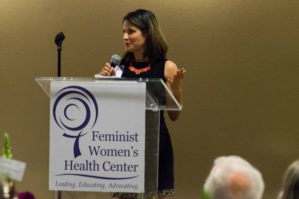 A staff member holding a microphone speaks from behind a podium bearing the Feminist Women’s Health Center logo.