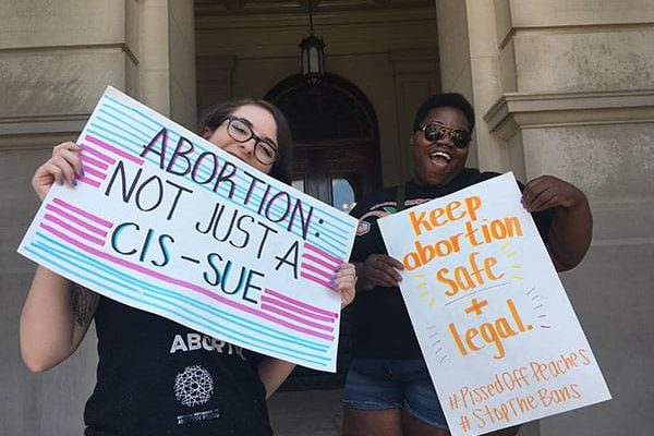 Two demonstrators pose with posters reading “Abortion is not a CIS-SUE” and “Keep Abortion Safe and Legal #PissedOffPeaches #StopTheBans” at a rally.